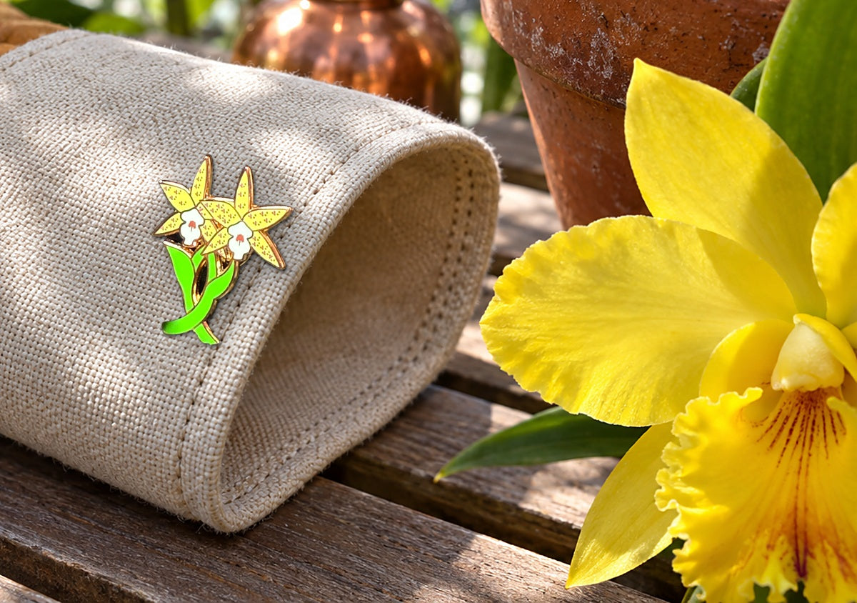 Yellow Double Laelia Cattleya orchid enamel pin on a canvas greenhouse glove cuff beside a yellow Cattleya bloom