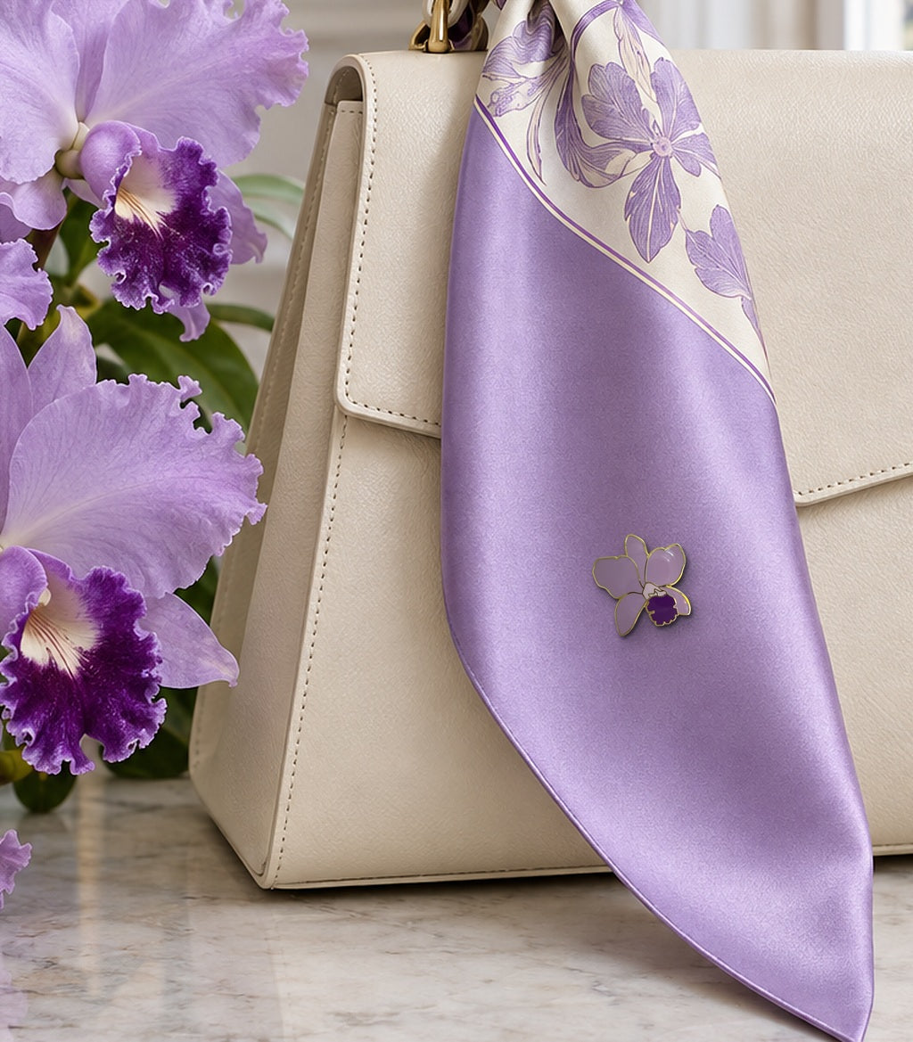 Lavender Lueddemannia orchid enamel pin on a lilac silk scarf tied to an ivory handbag beside lavender Cattleya blooms