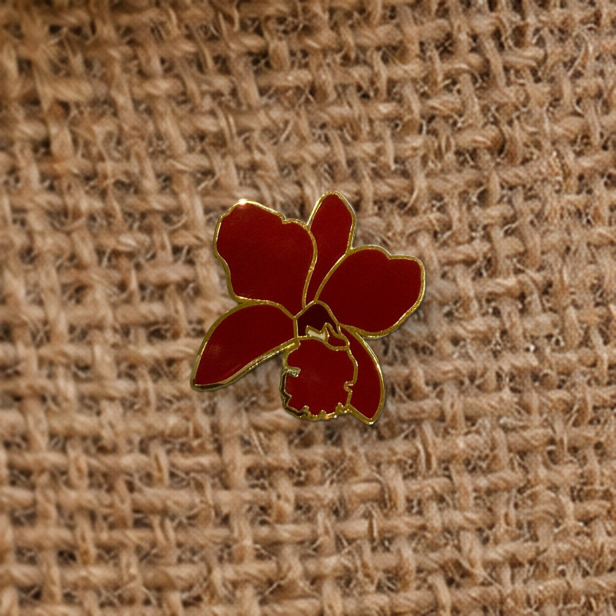 Close-up of the deep red Lueddemannia orchid enamel pin on burlap