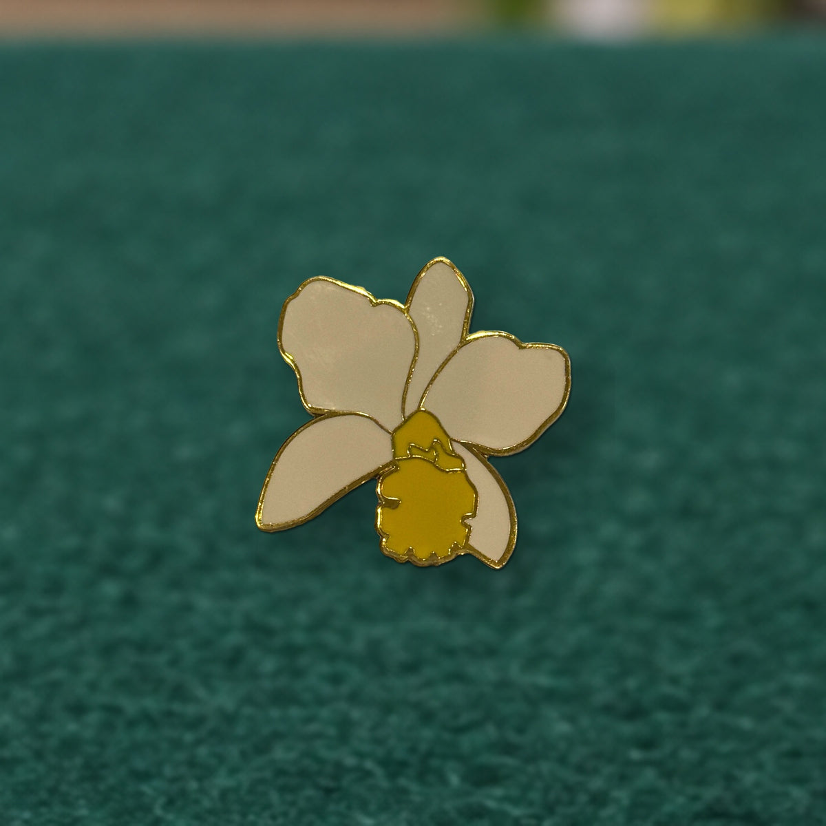 Close-up of the white and yellow Lueddemannia orchid enamel pin with gold outline on green felt
