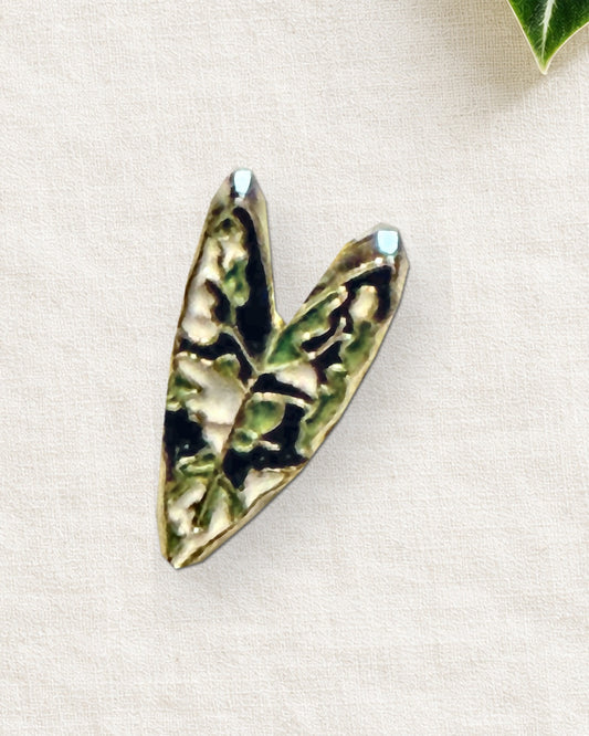 Close-up of the mini Alocasia Frydek enamel pin with green veins, dark velvet-leaf enamel and gold-tone outline on cream linen