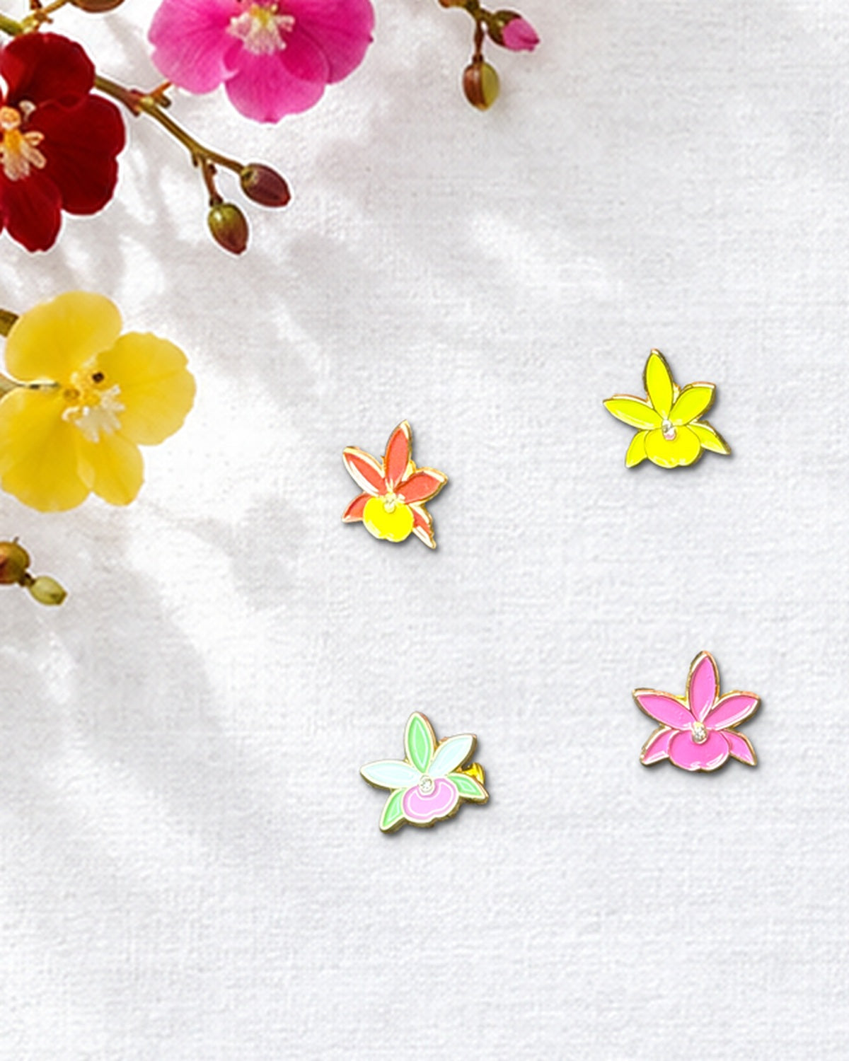 Red, Lime, Mixed Lime and Pink, and Fuchsia mini Broughtonia orchid enamel pins on white linen beside fresh Broughtonia blooms