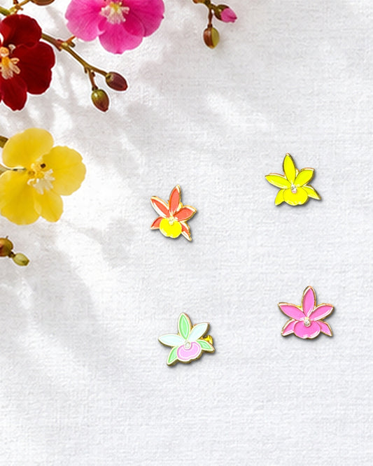 Red, Lime, Mixed Lime and Pink, and Fuchsia mini Broughtonia orchid enamel pins on white linen beside fresh Broughtonia blooms