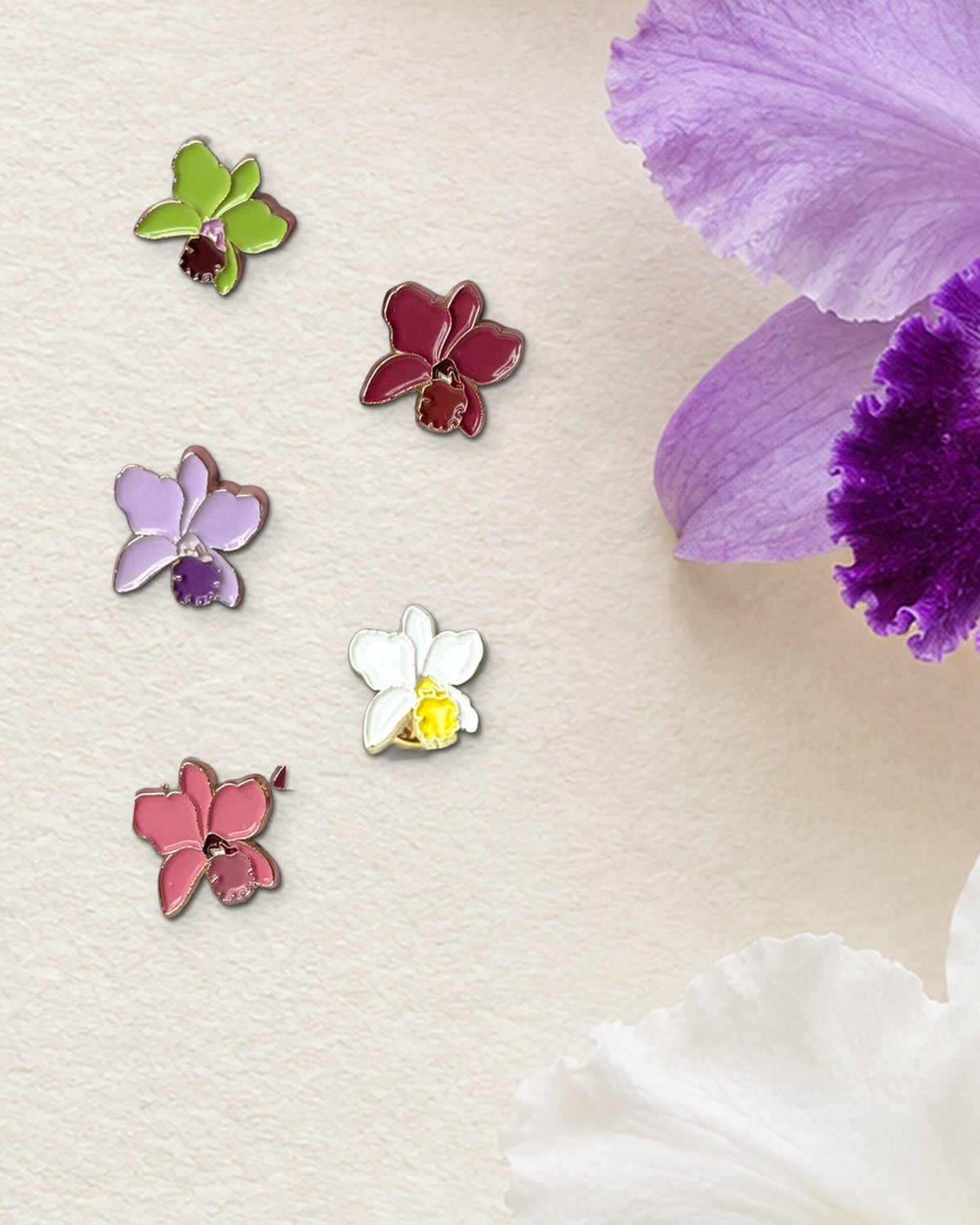 Five 14mm mini Cattleya orchid enamel pins in Green, Red, Purple, White and Pink on botanical watercolor paper beside fresh lavender and white Cattleya blooms