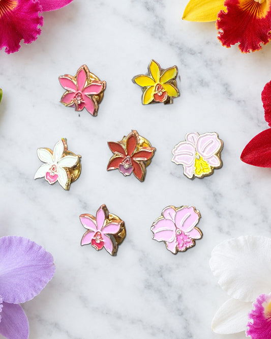 Seven 14mm mini Cattleya orchid enamel pins on white marble ringed by fresh pink, yellow, red, lavender and white Cattleya blooms