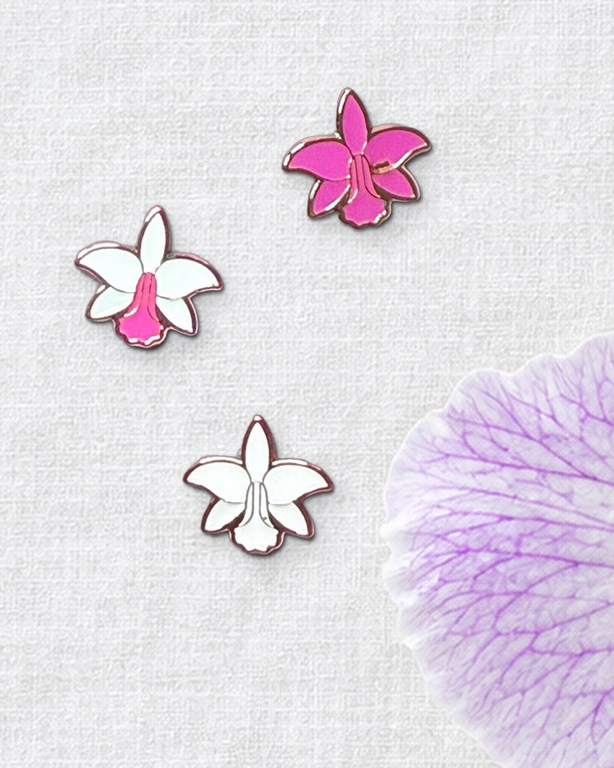 Three 14mm mini Cattleya violacea orchid enamel pins in White Pink, Pink and White on ivory linen beside a fresh violacea bloom