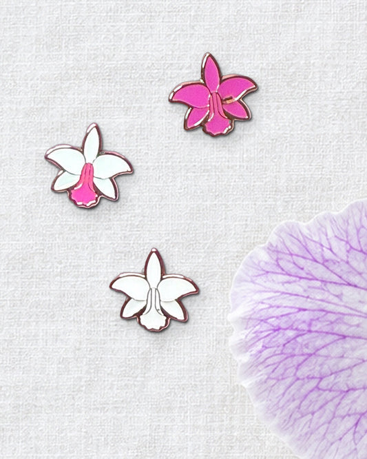 Three 14mm mini Cattleya violacea orchid enamel pins in White Pink, Pink and White on ivory linen beside a fresh violacea bloom