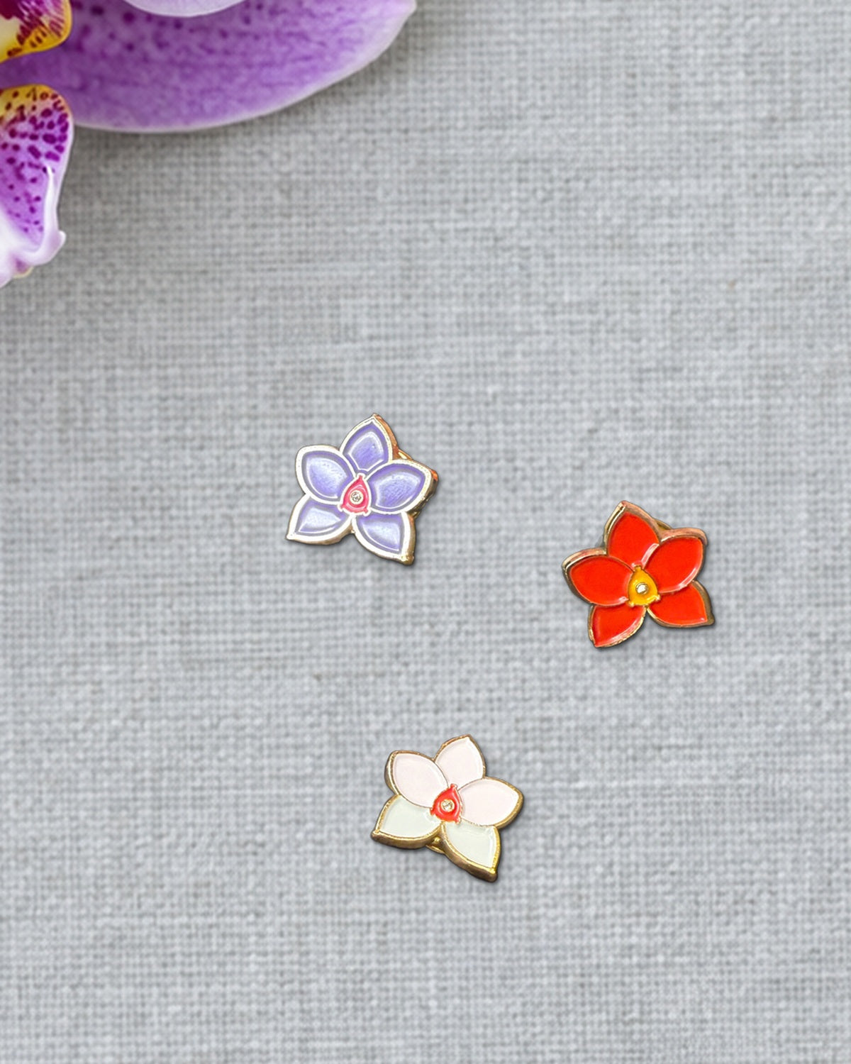 Purple, Red and Multi mini novelty Phalaenopsis enamel pins on grey linen beside a fresh spotted lavender Phalaenopsis bloom