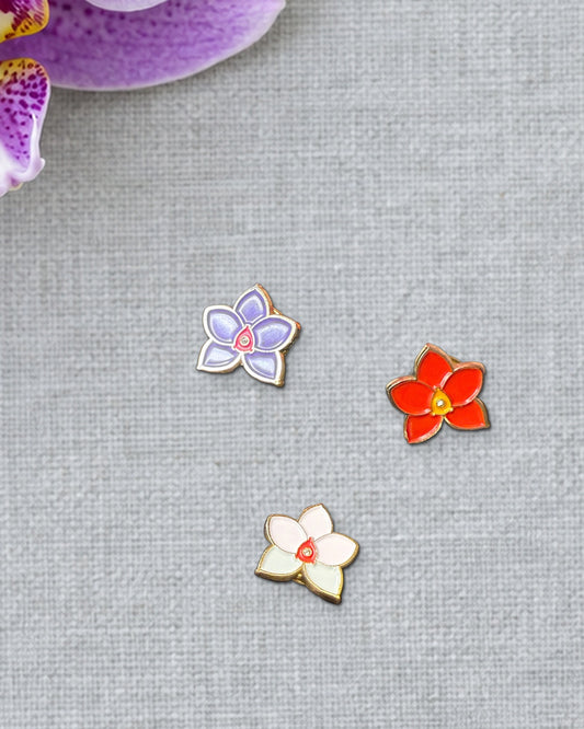 Purple, Red and Multi mini novelty Phalaenopsis enamel pins on grey linen beside a fresh spotted lavender Phalaenopsis bloom