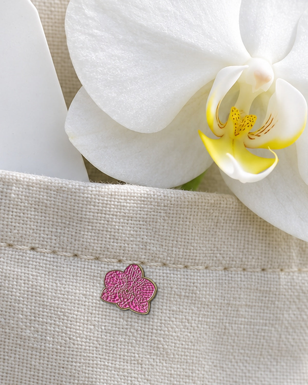Fuchsia mini Phalaenopsis enamel pin on a linen greenhouse apron pocket with a plant tag and a fresh white Phalaenopsis bloom