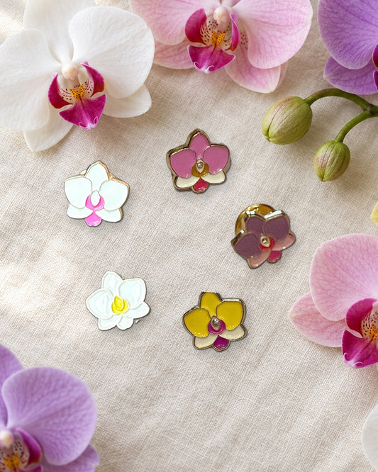 All five mini Phalaenopsis orchid enamel pins on ivory linen among fresh white, pink, lavender and yellow Phalaenopsis blooms and buds