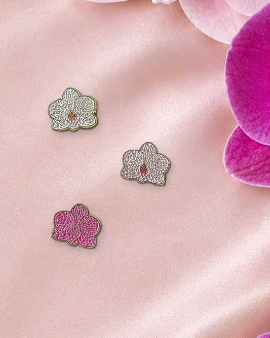 Three 14mm mini Phalaenopsis orchid enamel pins in White, Pink and Fuchsia on blush silk beside fresh pink and fuchsia Phalaenopsis blooms