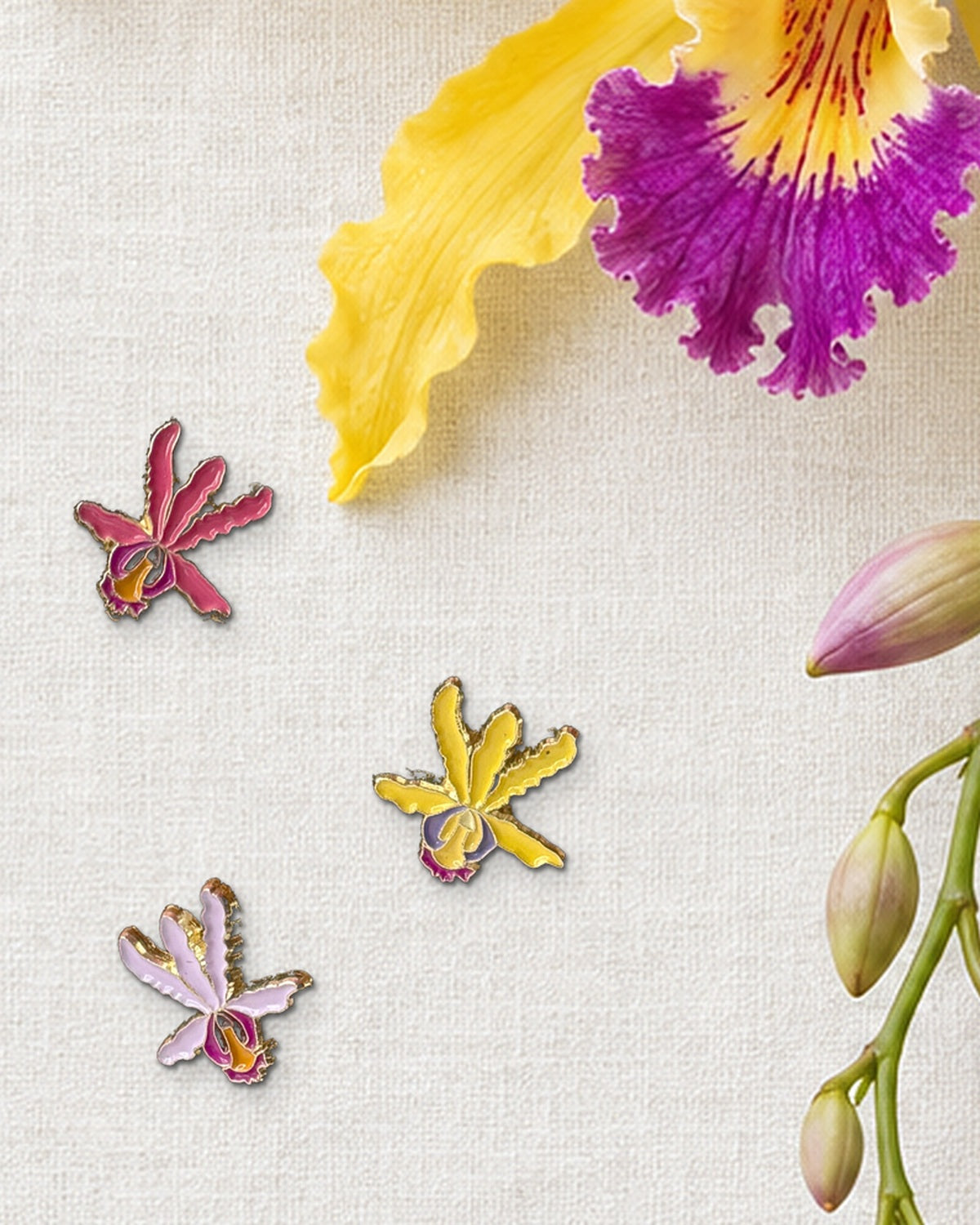 Three 14mm mini Schomburgkia orchid enamel pins in Rusty, Yellow and Pink Lavender on ivory linen beside a fresh yellow orchid bloom and buds