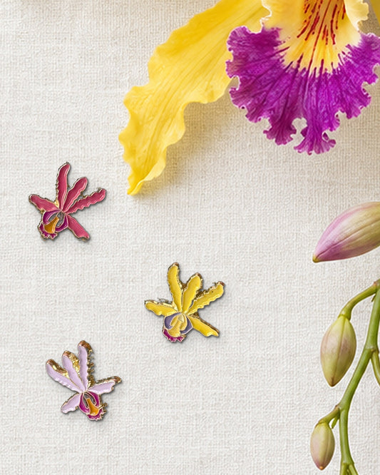 Three 14mm mini Schomburgkia orchid enamel pins in Rusty, Yellow and Pink Lavender on ivory linen beside a fresh yellow orchid bloom and buds