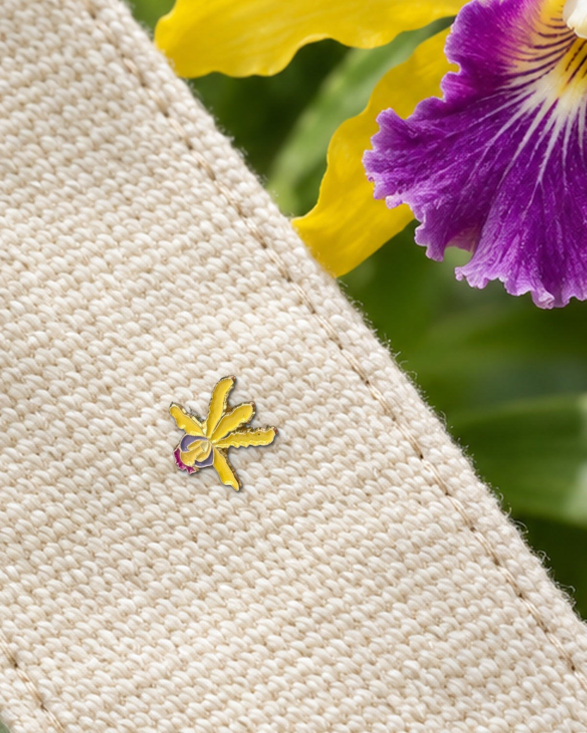 Yellow mini Schomburgkia enamel pin on a canvas bag strap beside a real yellow orchid with a purple lip in the garden