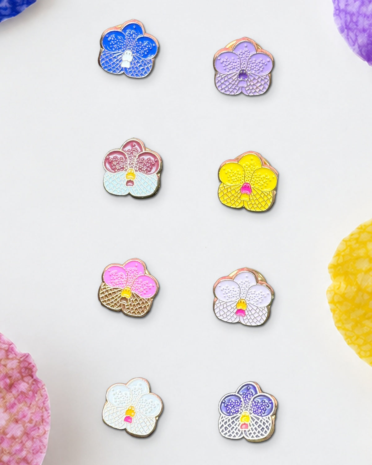 All eight 14mm mini Vanda orchid enamel pins on a white ceramic tray framed by fresh checkered blue, purple, yellow and pink Vanda blooms