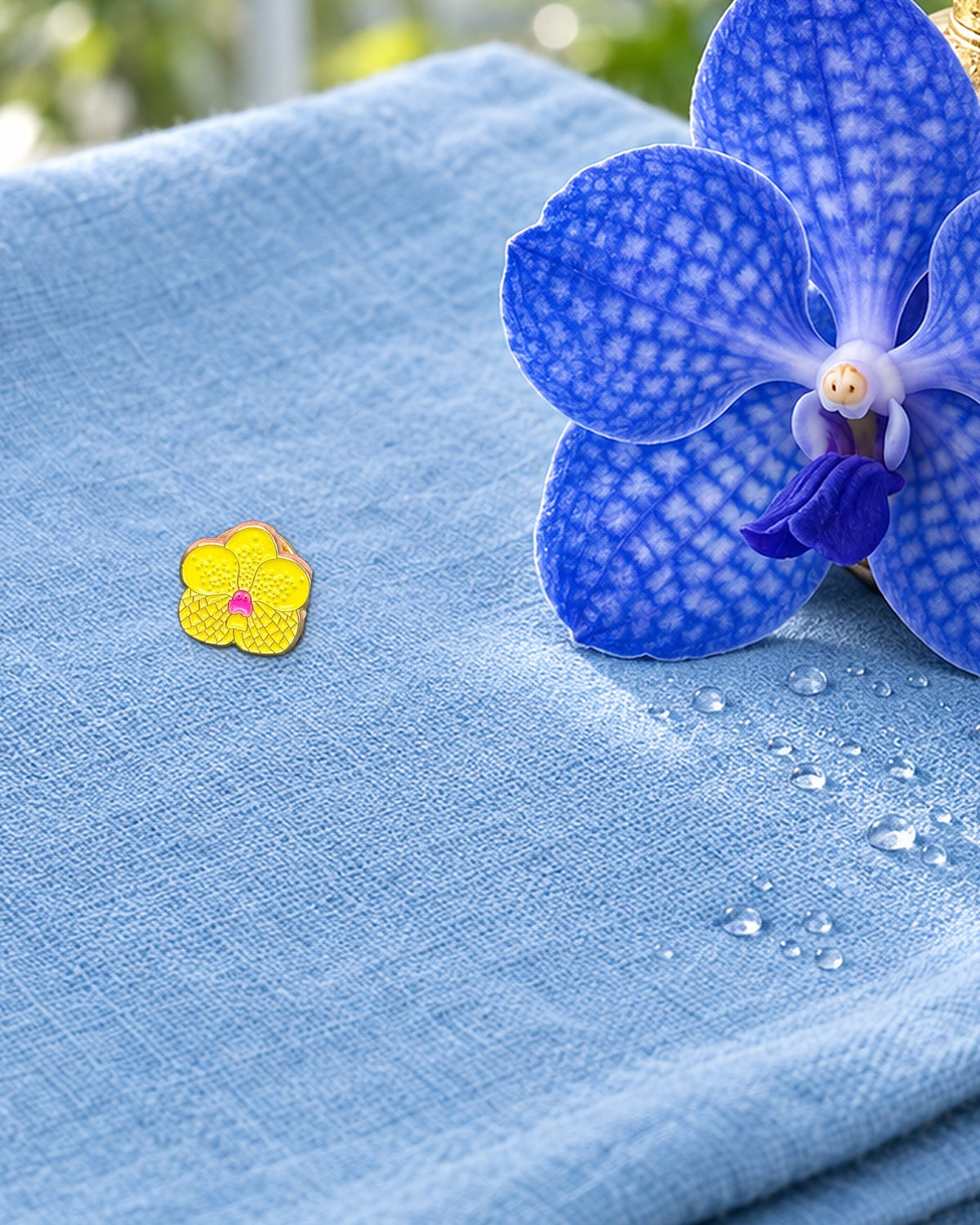 Yellow mini Vanda enamel pin on a sky-blue linen towel beside a brass plant mister and a fresh blue Vanda bloom