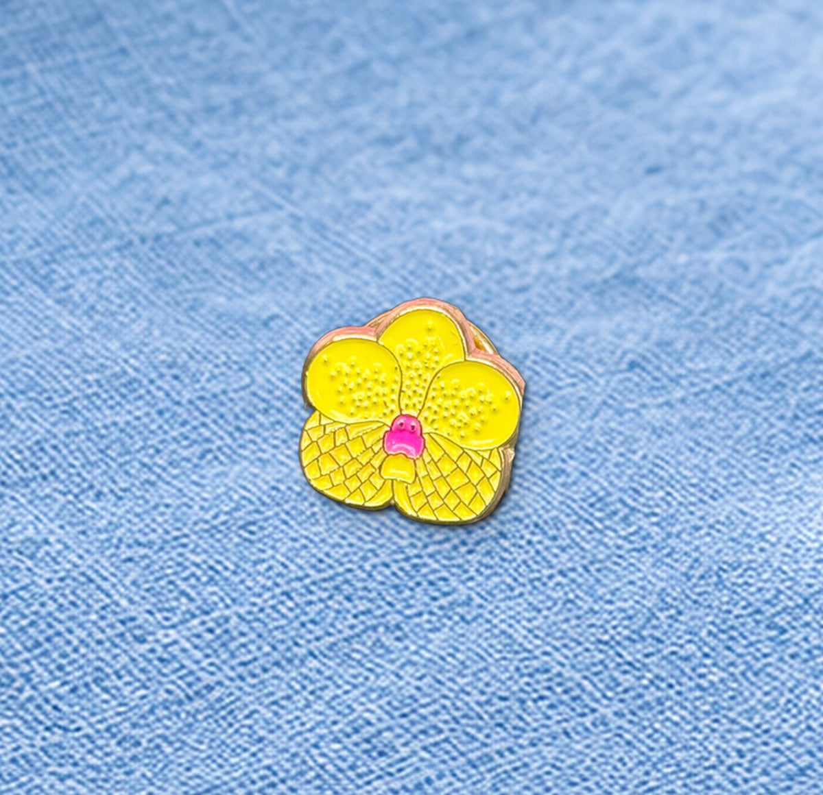 Close-up of the Yellow mini Vanda enamel pin with checkered petal texture and gold-tone outline on blue linen