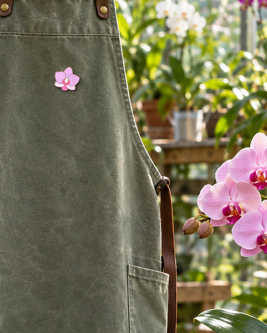 Pink Phal Cutie orchid enamel pin on a sage canvas greenhouse apron beside a blooming pink Phalaenopsis