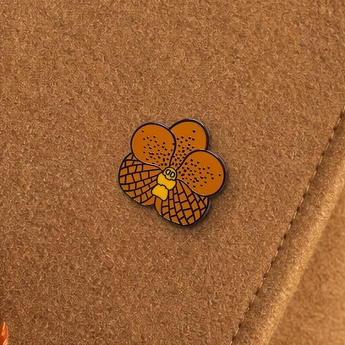 Orange Vanda enamel pin close-up on camel wool